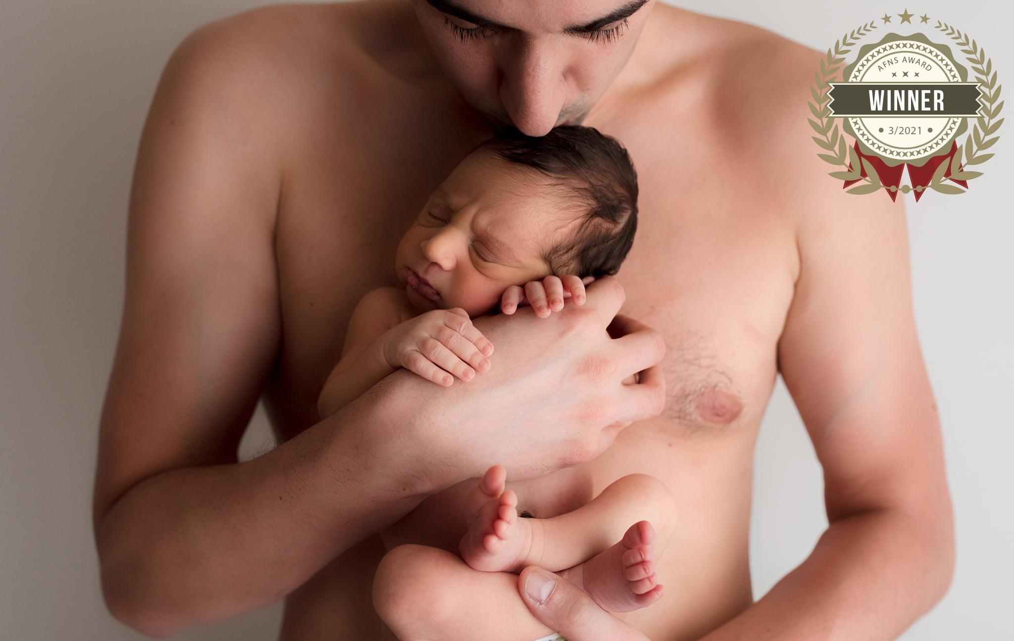 Shirtless dad holding newborn and kissing him on the head with award badge on top right.