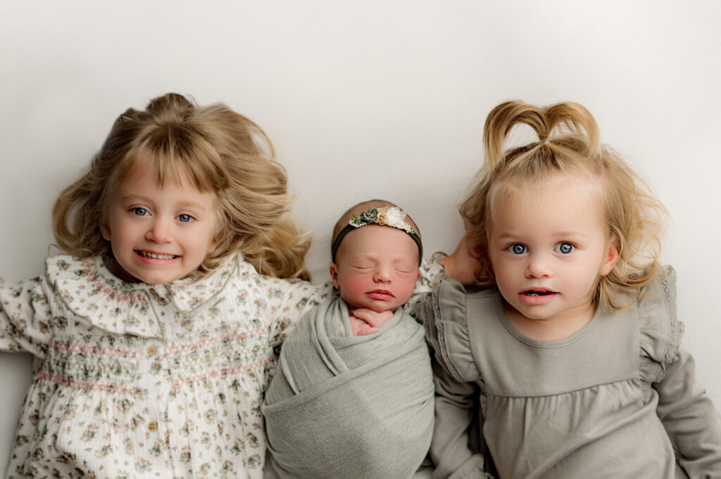 Two older sisters looking a the camera while their newborn sister is between them for her charlotte newborn photoshoot