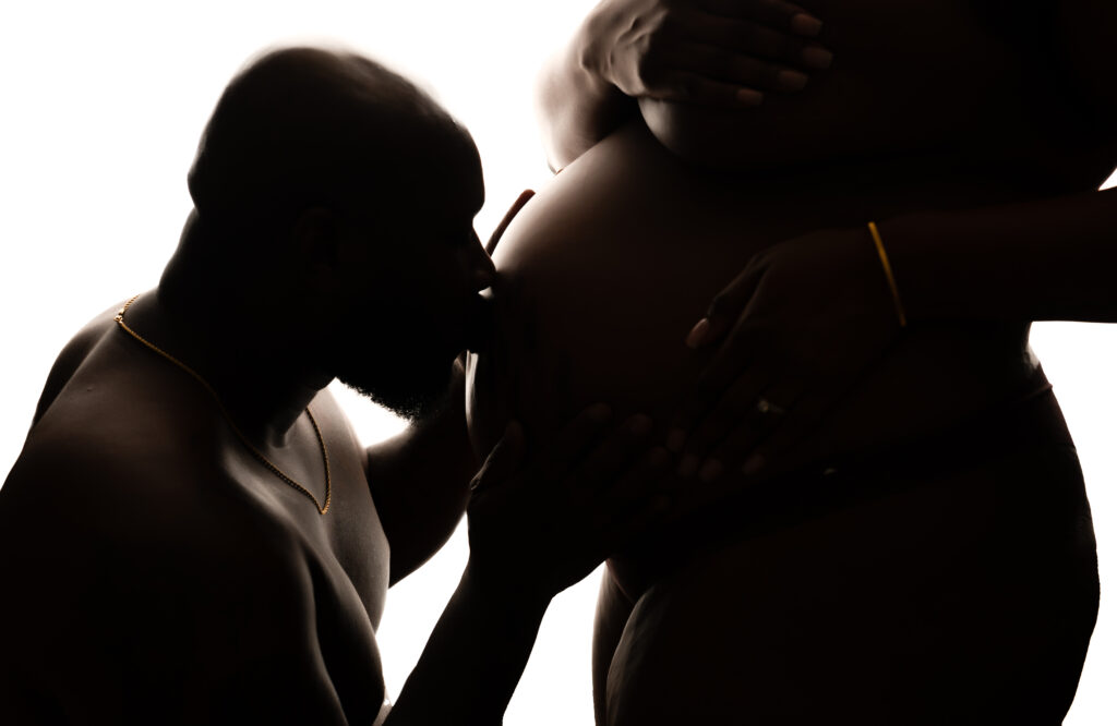 Shirtless dad kissing the exposed pregnant belly of his wife during a charlotte maternity boudoir session