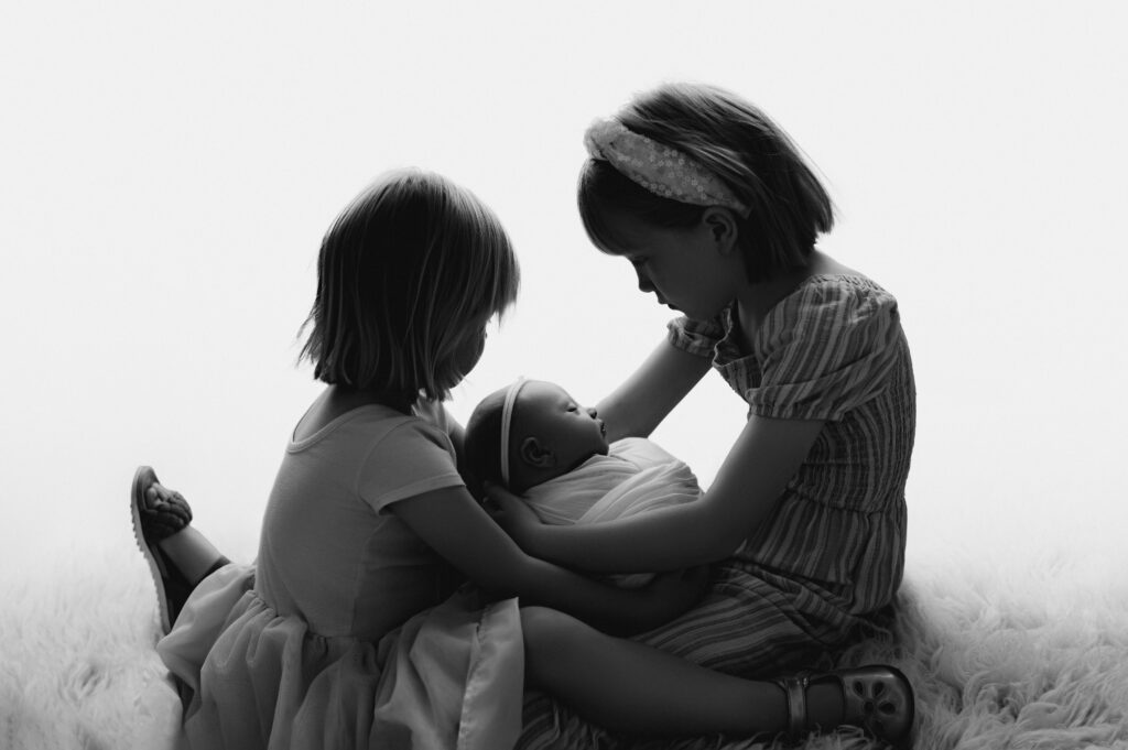 Two older sisters facing each other while holding and looking down at their newborn baby sister at a charlotte newborn photography studio