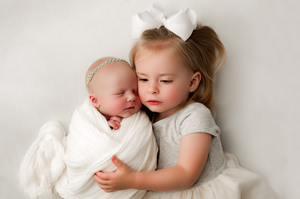 Toddler with her arms wrapped around baby sister with the newborn and sisters face pressed up against each other 