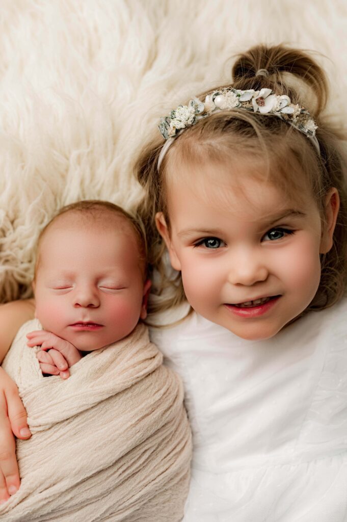 Little girl with her newborn brother laying down smiling and looking into the camera for his professional newborn photography session