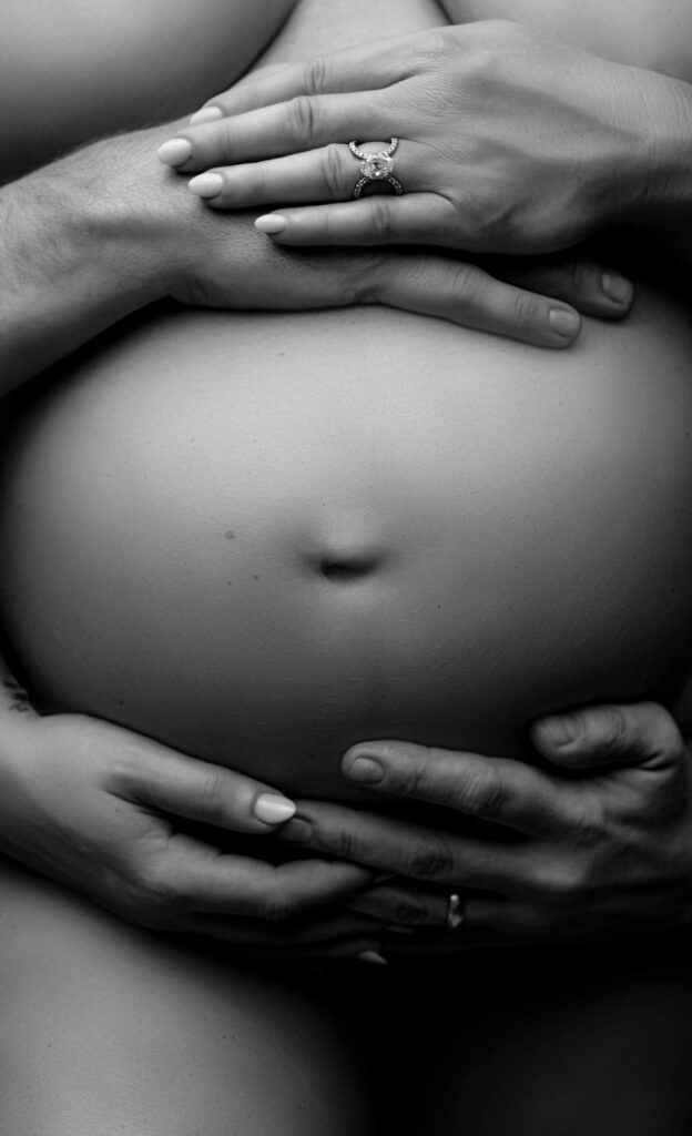 Black and white photo of pregnant belly with the hands of the mother and father surrounding it 