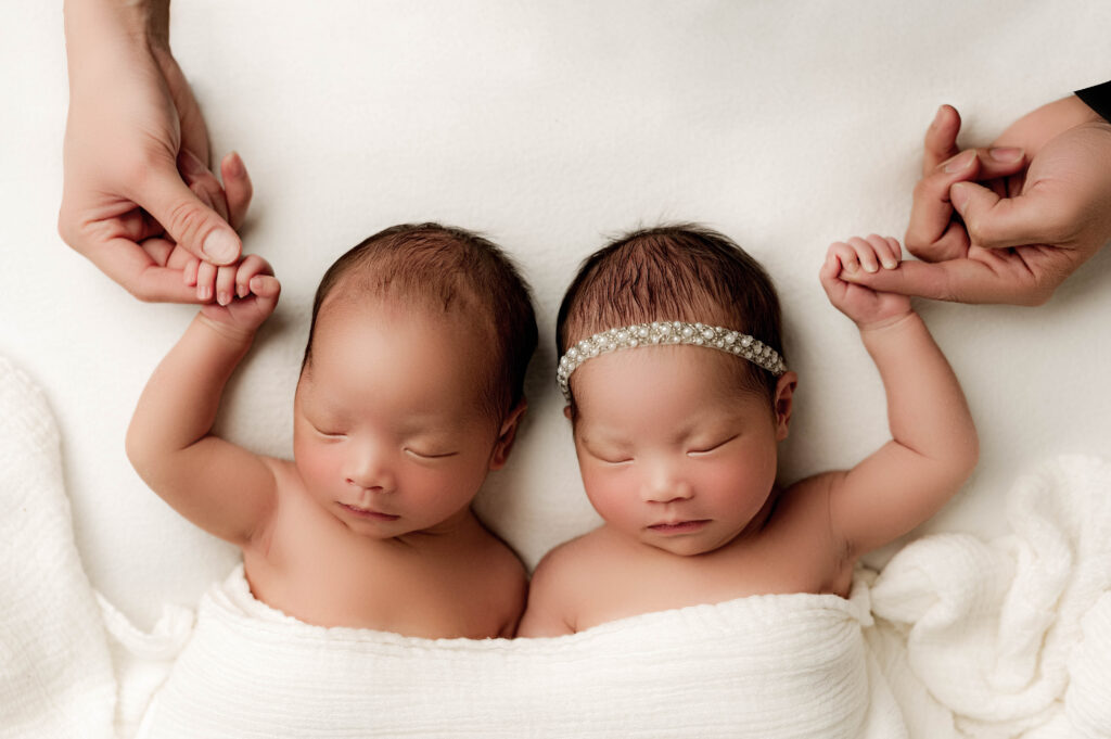 Twin newborns laying on their back sleeping while each baby holds the hand of their parents during charlotte newborn photoshoot.