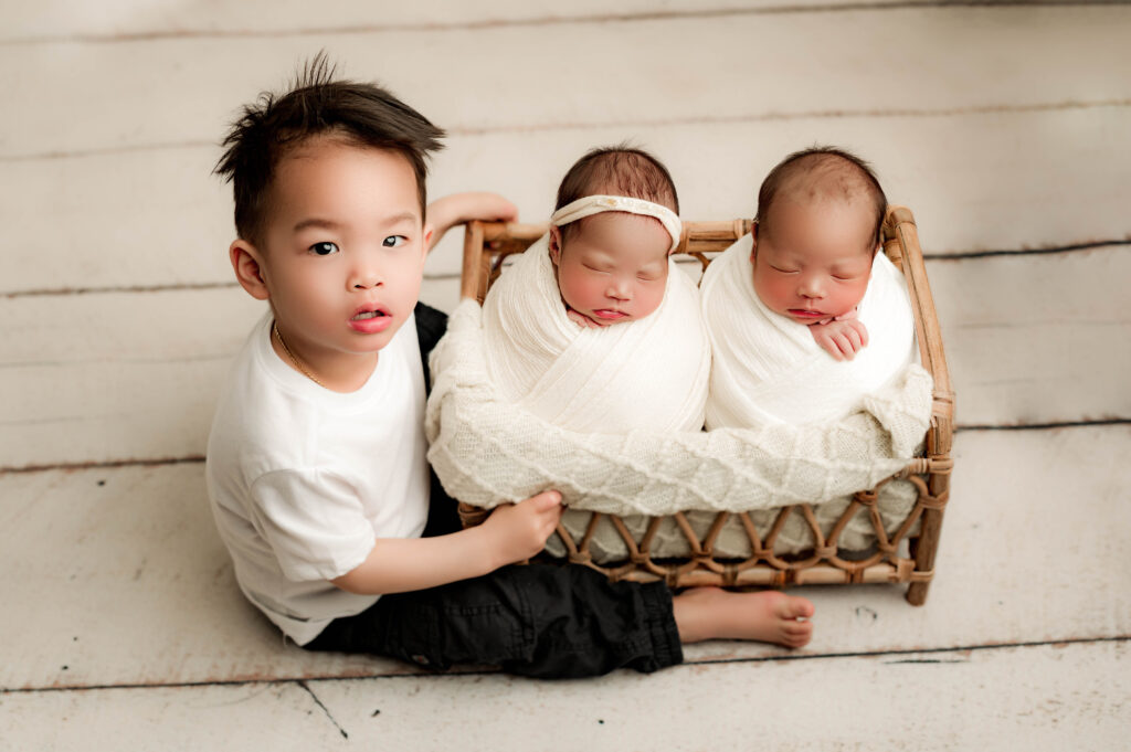 Big brother holding the bed with his twin brother and sister in it posing for their newborn photos.