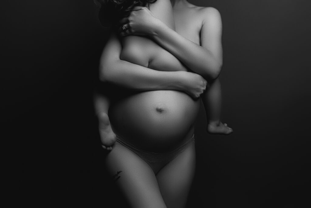 Black and white photo of mother holding her child on her pregnant belly showcasing her curves and raw beauty