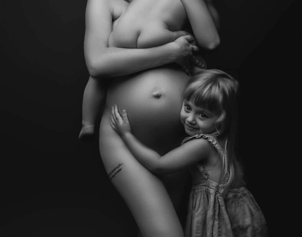 Black and white photo of the same mother holding her child on her pregnant belly while her oldest daughter hugs her belly taken at her second maternity session showcasing her curves and raw beauty