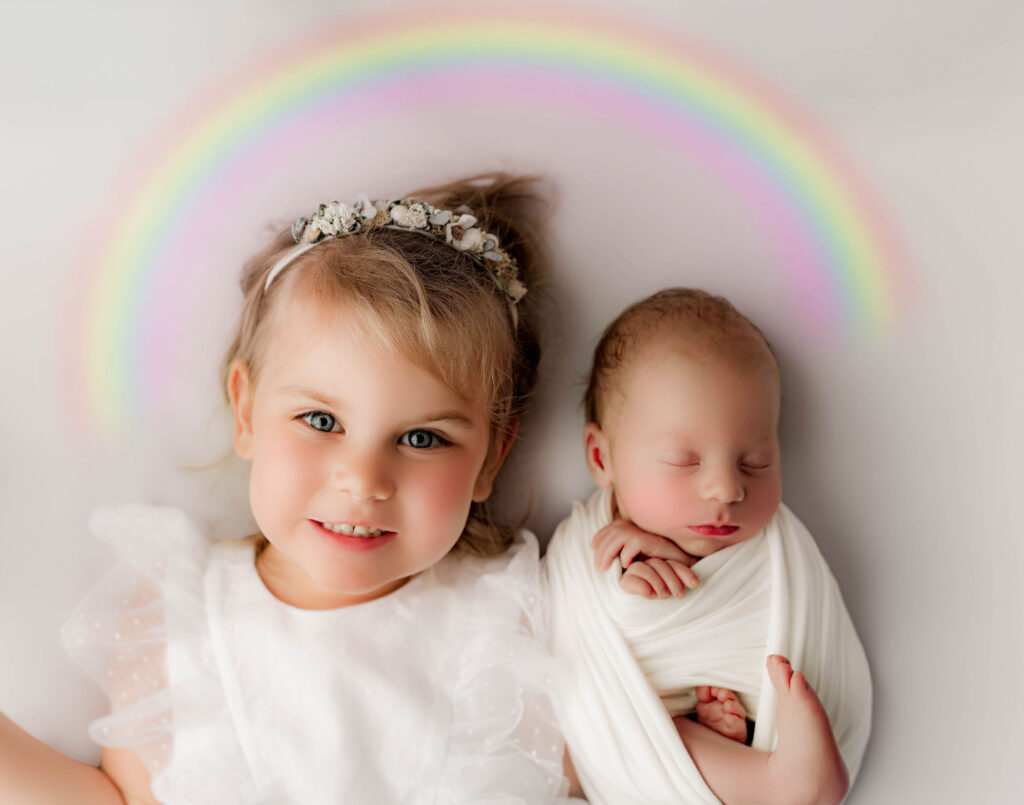 Toddler next to her newborn brother laying down with a rainbow over their heads to show they are rainbow babies during newborn photography session at a charlotte photography studio