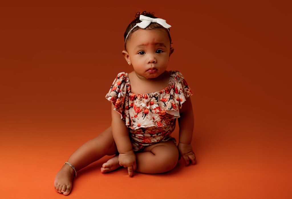 Little girl with pout face sitting on orange backdrop pointing at the ground while looking into the camera. 