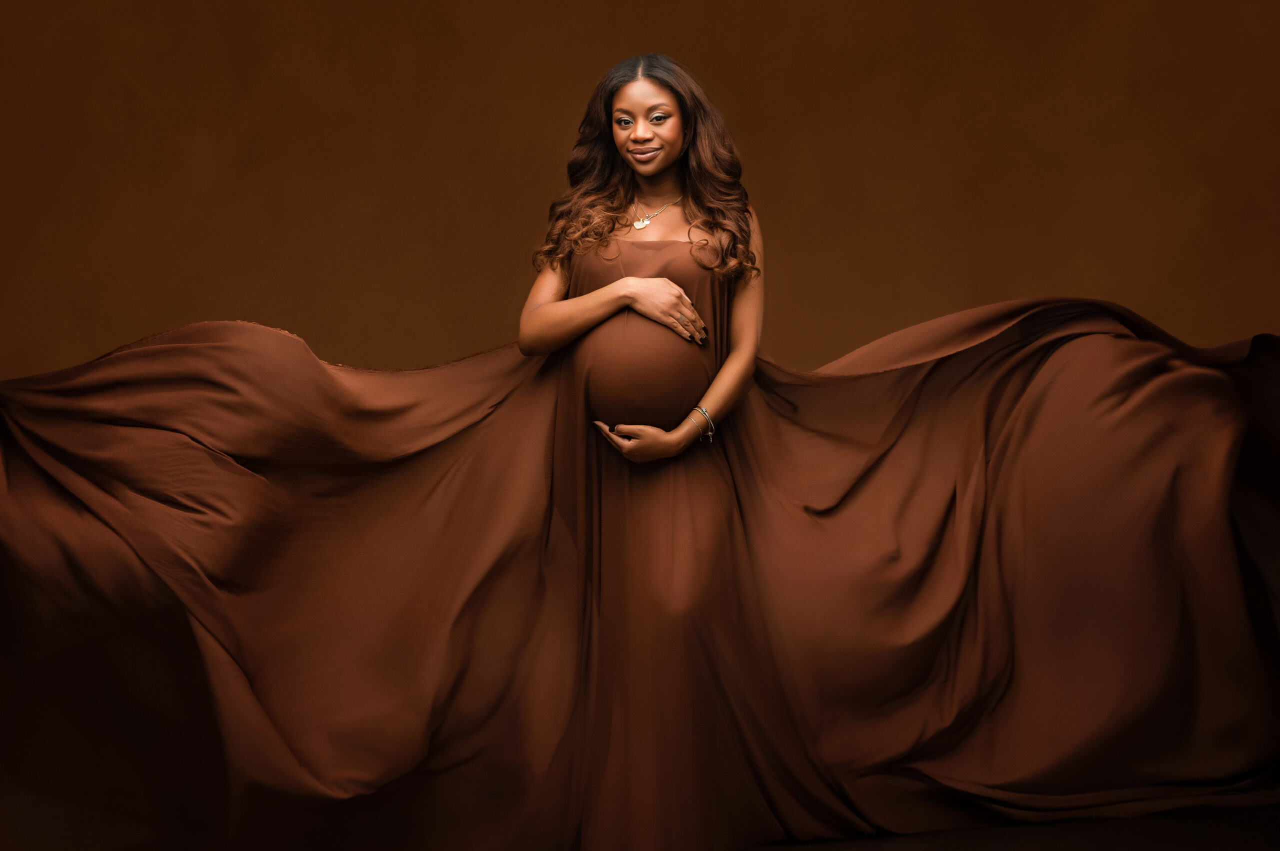 Pregnant women looking at camera holding her belly while brown fabric flows beside her..