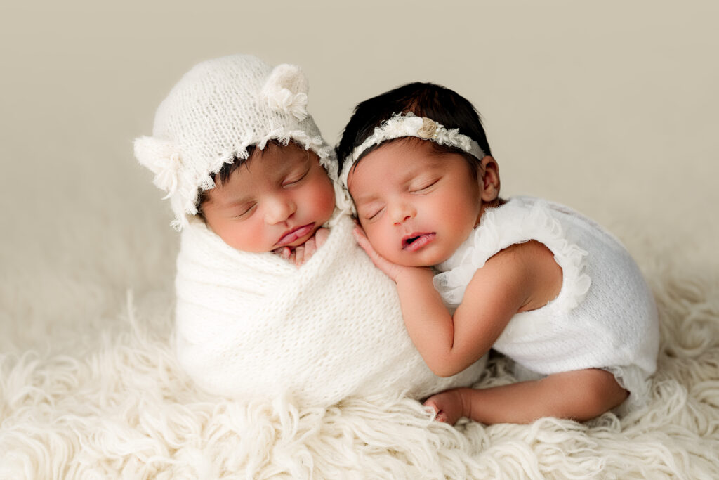 Twin girls asleep one with wrapped up in a "potato wrap" and bear bonnet on with bows and the other one leaning against her sister wearing a white outfit with ruffles on her shoulders. 