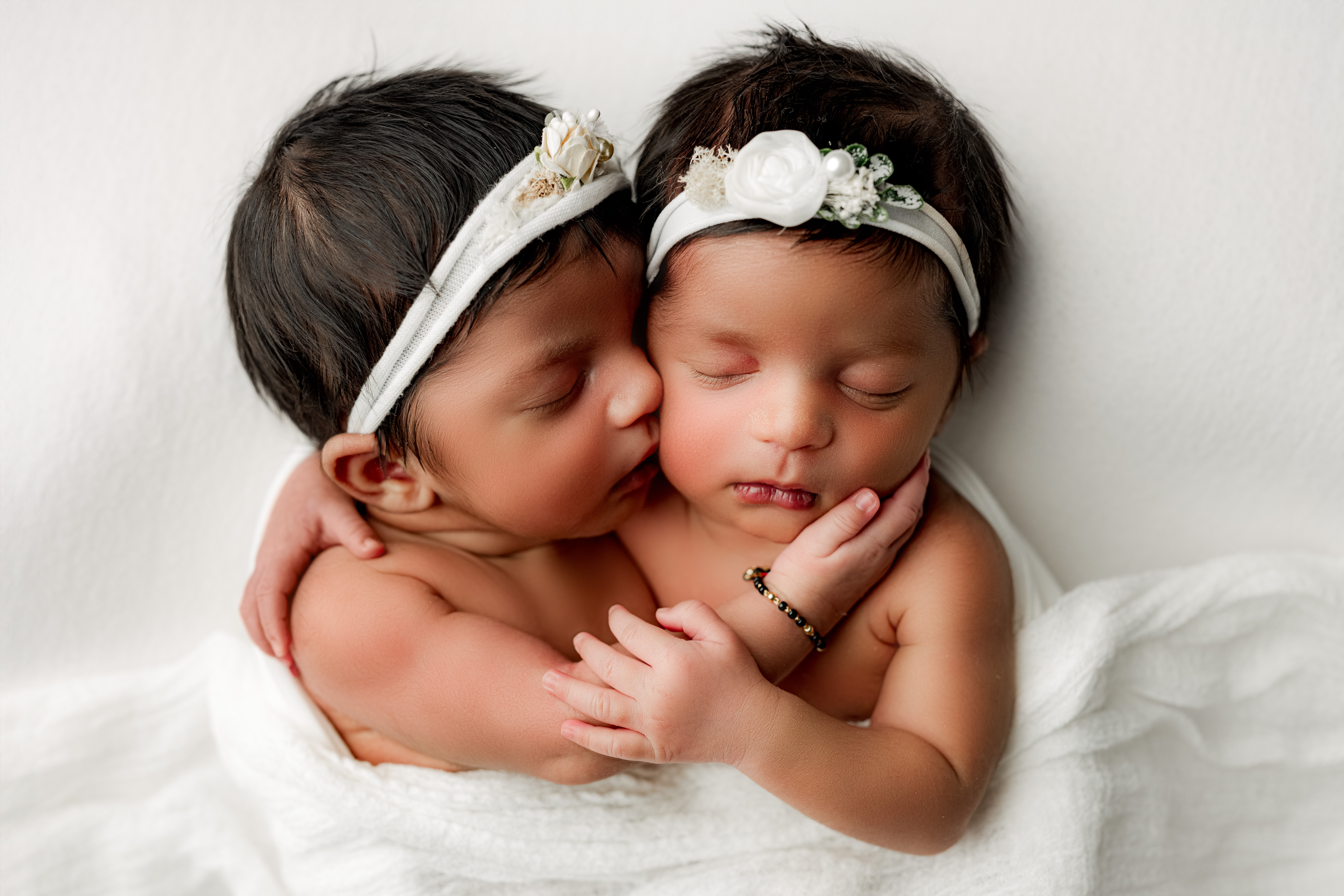 Twin girls on white backdrop with one baby kissing her sisters cheek while her sister cuddles her during Charlotte Newborn Photography session