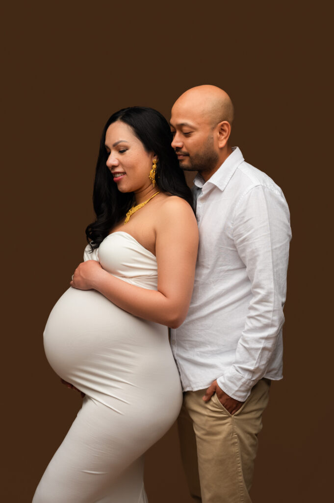 Studio maternity photo of a couple in soft, romantic lighting, with the partner standing closely behind the mother-to-be, gently resting his hand on her belly—captured during a Charlotte maternity photography session.