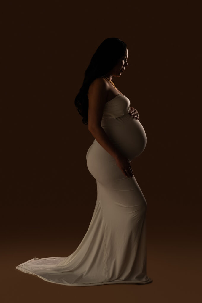Silhouetted side profile of pregnant woman in fitted white gown, elegant studio lighting, during her third trimester.