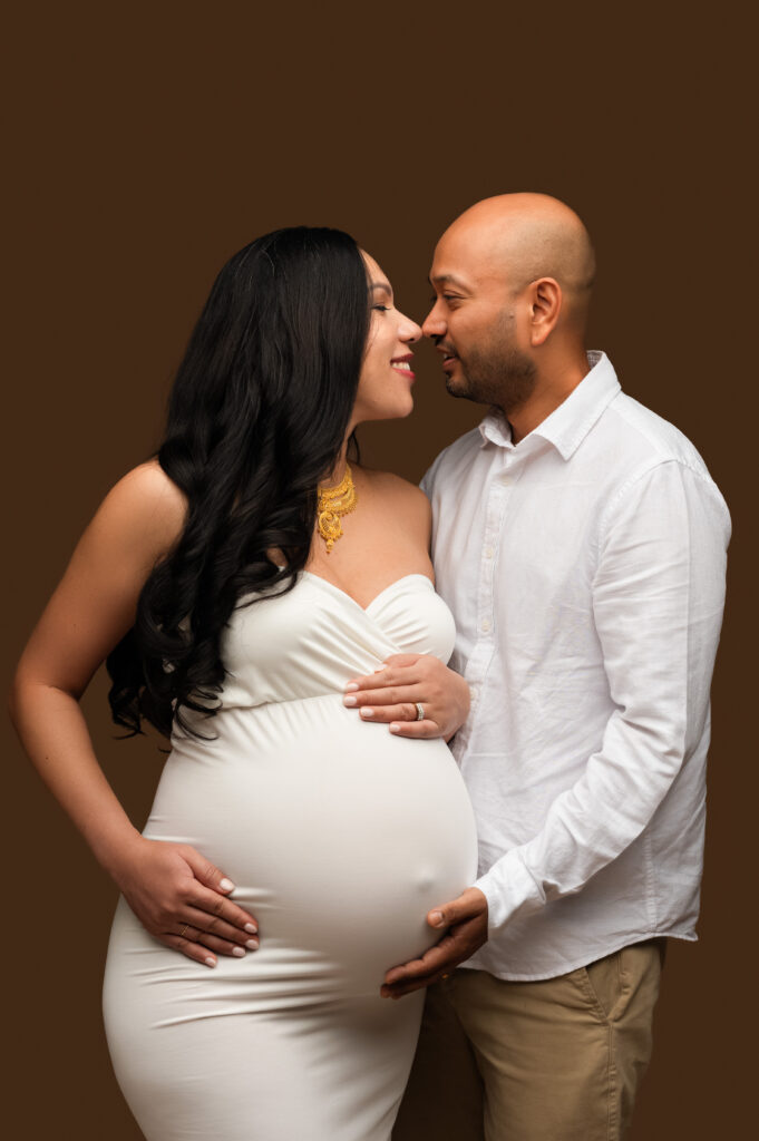 Maternity portrait of an expectant couple standing face to face and smiling, with the father-to-be gently holding the baby bump, taken at a Charlotte maternity studio by NicSo Studio.