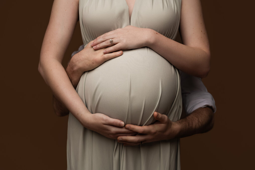 Close-up photo of a pregnant belly embraced by both parents’ hands, with delicate textures of a neutral gown and wedding rings visible, during a Charlotte maternity studio session focused on connection.