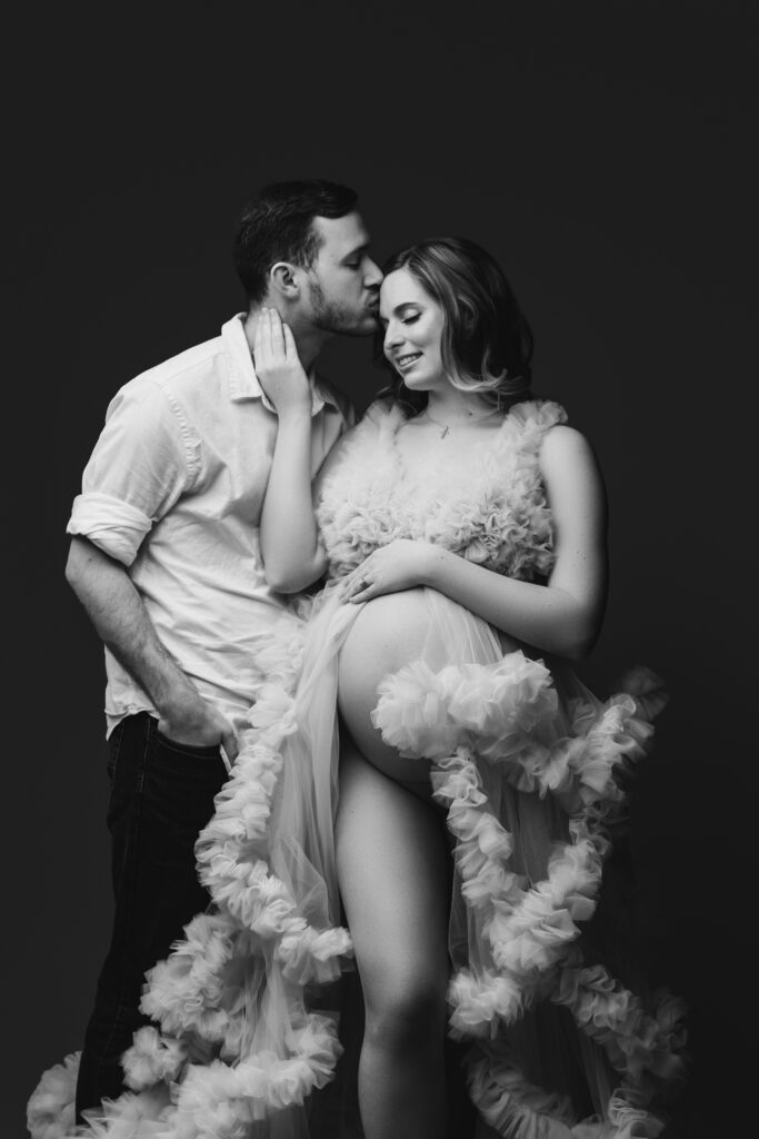 Black-and-white fine art maternity portrait of a couple sharing a joyful moment, with the partner gently kissing the mother-to-be’s forehead, photographed at a luxury Charlotte maternity photography studio.