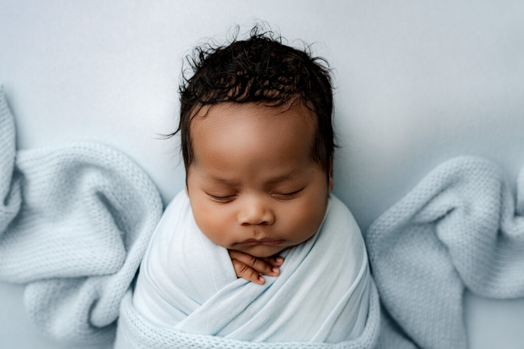 Swaddled newborn boy in soft blue wrap napping on pale blue backdrop taken at NicSo Studio, Charlotte, NC.