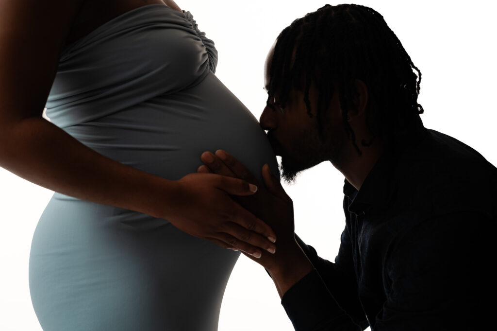 Silhouetted maternity portrait of a partner kneeling to kiss the baby bump, highlighting love and tenderness during a modern maternity session in-studio.
