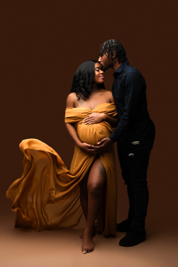 Studio maternity portrait of a pregnant woman in a flowing mustard gown with her partner gently kissing her forehead, styled and photographed in a luxury Charlotte maternity studio.

