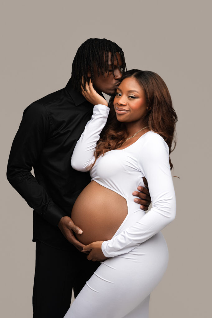 Stylish maternity photo of a couple in coordinated white and black outfits, with the partner holding the baby bump and the mother-to-be gently touching his face.