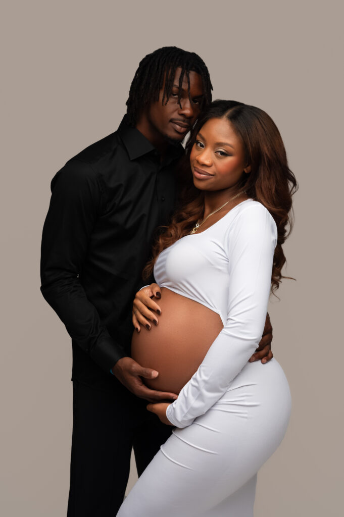 Romantic studio portrait of a pregnant couple, with the partner both mom to be and her partner smiling softly at the camera, wearing a white gown.