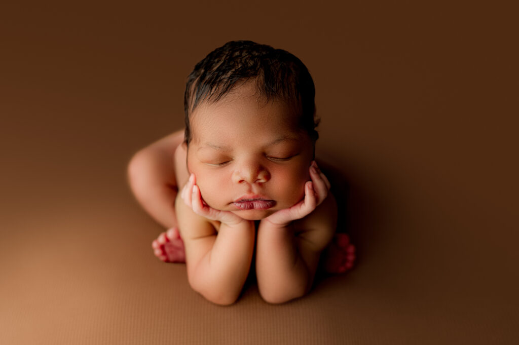Newborn in classic froggy pose, hands under chin on brown set, captured in Charlotte newborn photography studio.