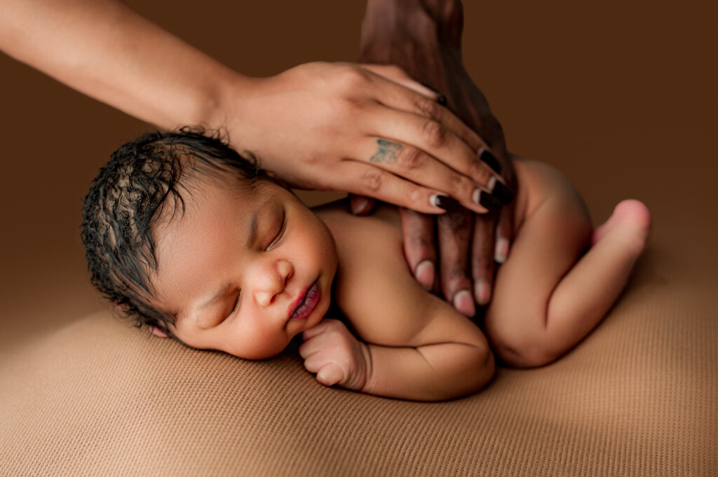 Parent’s hands resting gently on sleeping newborn posed on brown blanket during newborn session.

