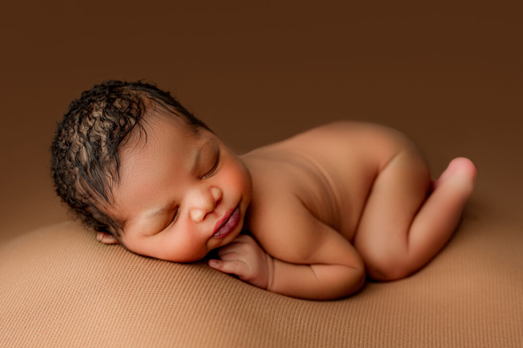 Newborn baby boy sleeping curled on tummy against warm brown backdrop.