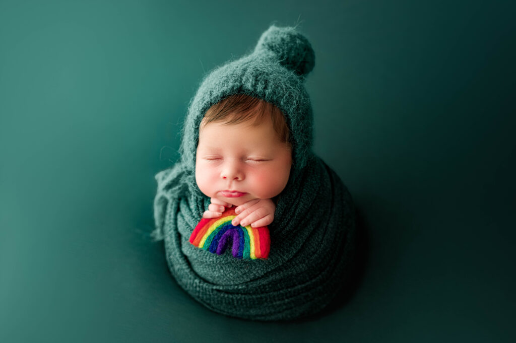 Newborn baby wrapped in deep green with matching bonnet, gently holding a small felt rainbow symbolizing hope after loss. Studio portrait by Charlotte rainbow baby photographer NicSo Studio.