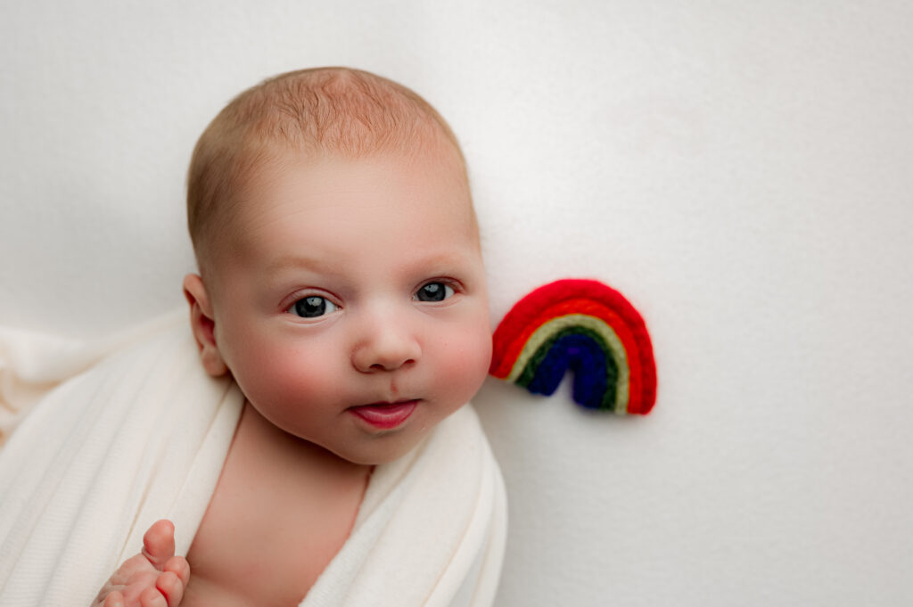 Alert newborn baby swaddled in white, gazing toward the camera with a small felt rainbow placed beside their cheek. Captured in Charlotte, NC by NicSo Studio to honor a rainbow baby journey.