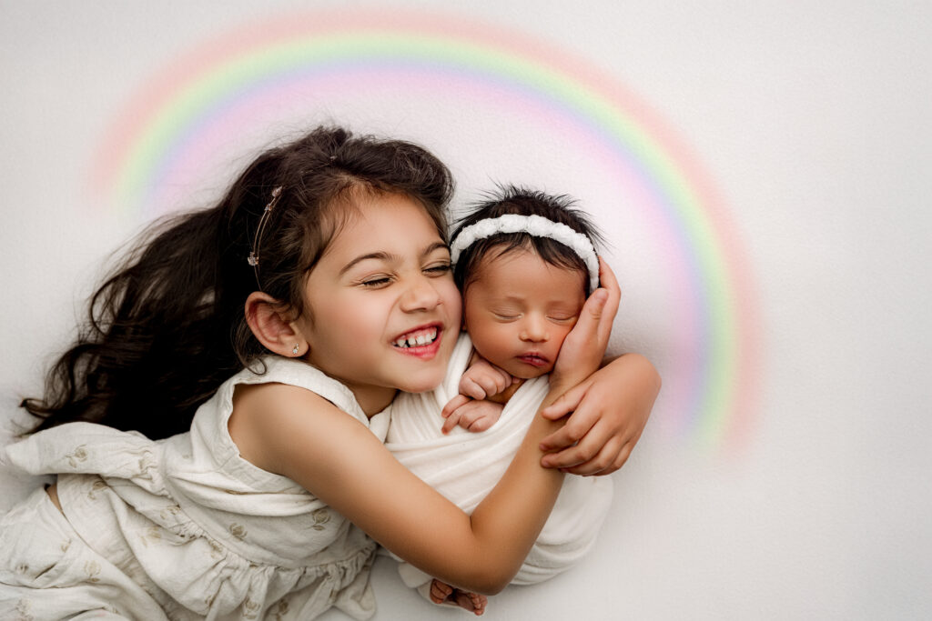 Joyful big sister lovingly cuddles her newborn rainbow baby sibling, both wrapped in white with a soft pastel rainbow digitally added above them. Both Rainbow Babies via frozen embryo IVF born 4 years apart. A sweet and emotional sibling portrait from NicSo Studio in Charlotte, NC.