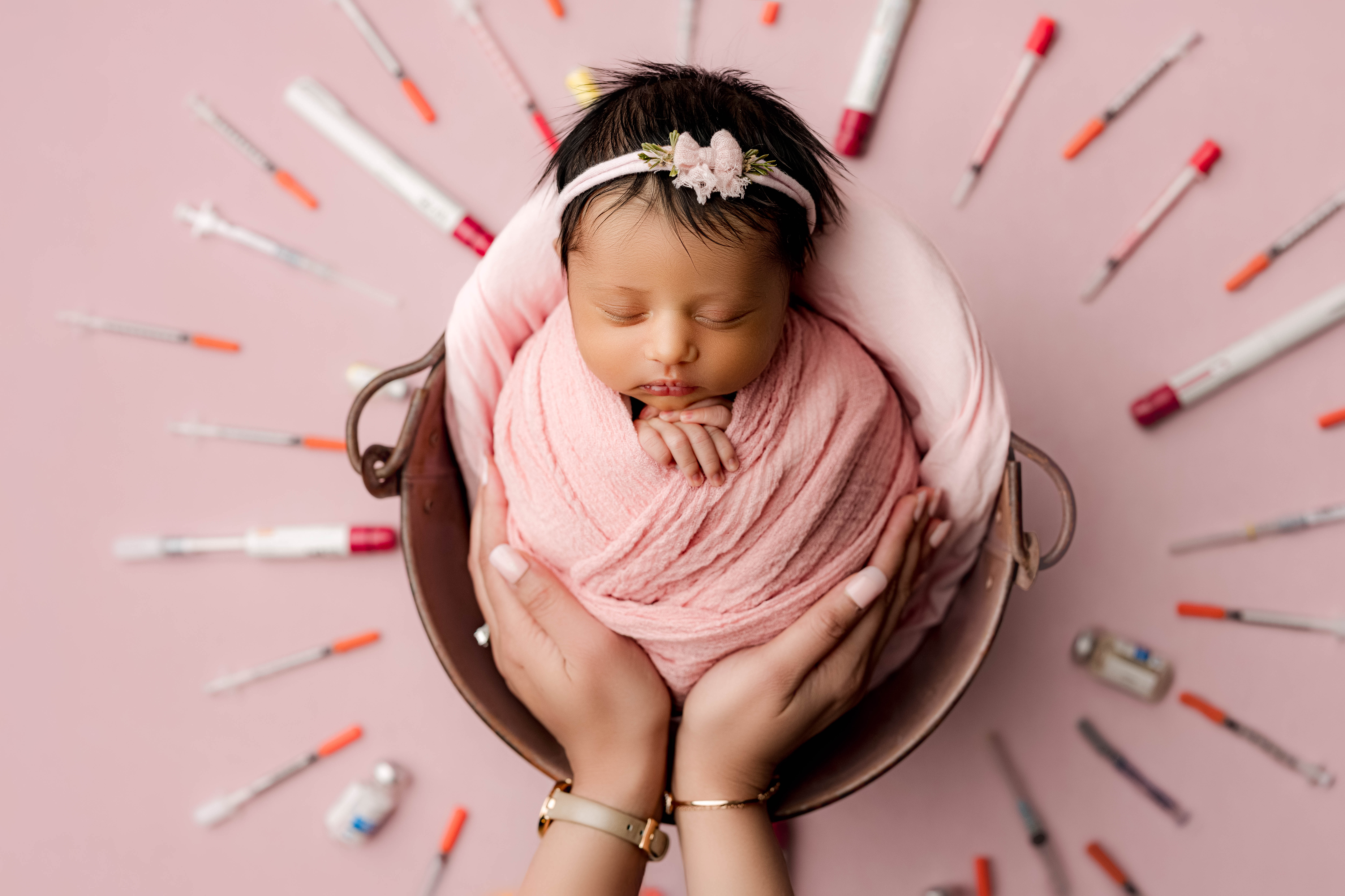 Newborn baby girl swaddled in soft pink wrap, peacefully sleeping in a basket, surrounded by IVF needles and medication. A powerful tribute to her parents’ infertility journey.