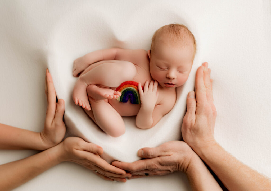 Newborn baby sleeping on soft white fabric, posed inside of a heart with a felt rainbow on their chest and gently cradled by their parents' hands. A heartfelt image honoring a rainbow baby’s journey, photographed at NicSo Studio in Charlotte.