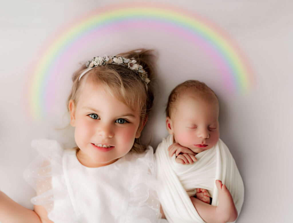 Young girl smiles while lying beside her sleeping newborn baby brother sibling, both under a pastel rainbow illustration celebrating a rainbow baby's arrival. Charlotte sibling and newborn portrait by NicSo Studio.
