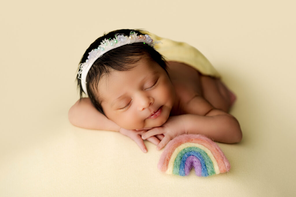 Newborn baby girl sleeping on yellow blanket with rainbow prop with a little smile during Charlotte rainbow baby session