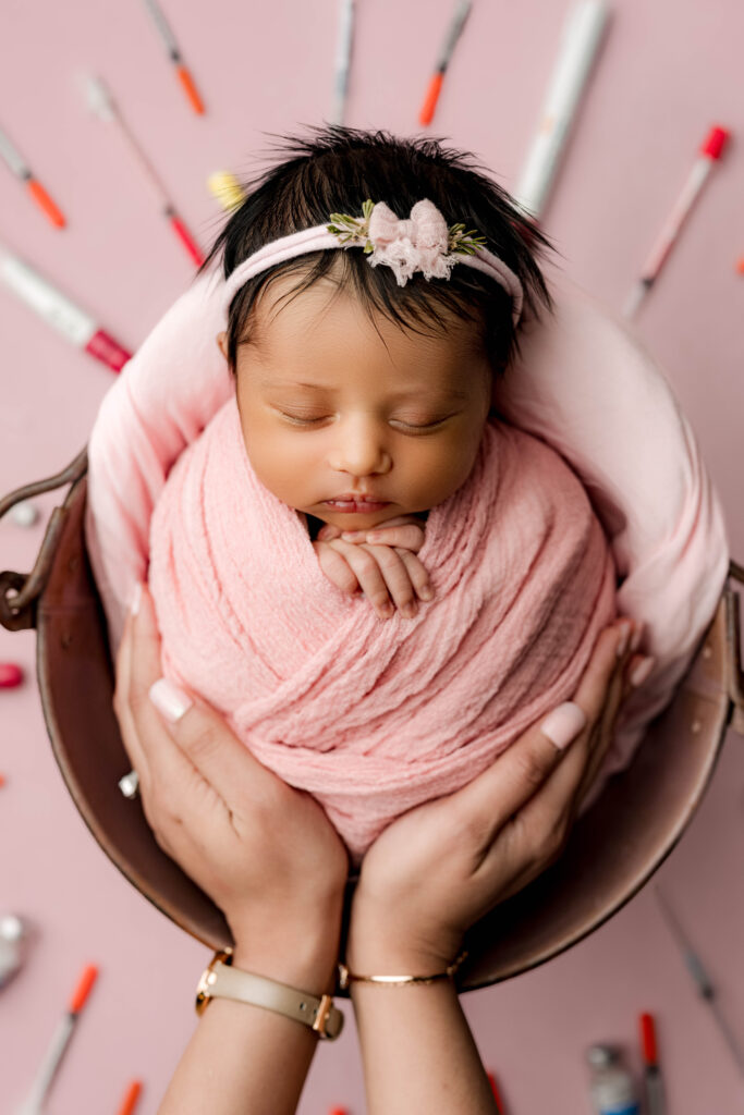 Newborn rainbow baby swaddled in pink surrounded by IVF and fertility medication syringes while being held by her mom's hands in tribute image