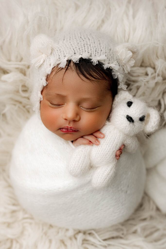 Newborn rainbow baby girl wrapped in white, holding a tiny teddy bear, resting on soft fur