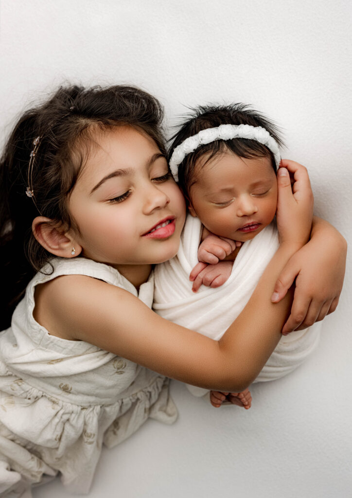 Big sister gently embracing newborn baby sister wrapped in white on matching white backdrop during Charlotte NC newborn photography session