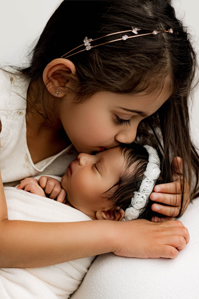 Close-up of big sister gently kissing swaddled newborn on white backdrop