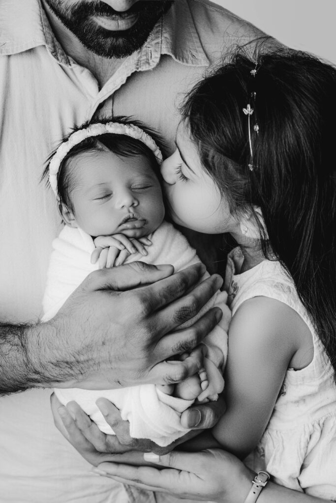 Big sister kissing newborn rainbow baby’s forehead while dad gently cradles them both
