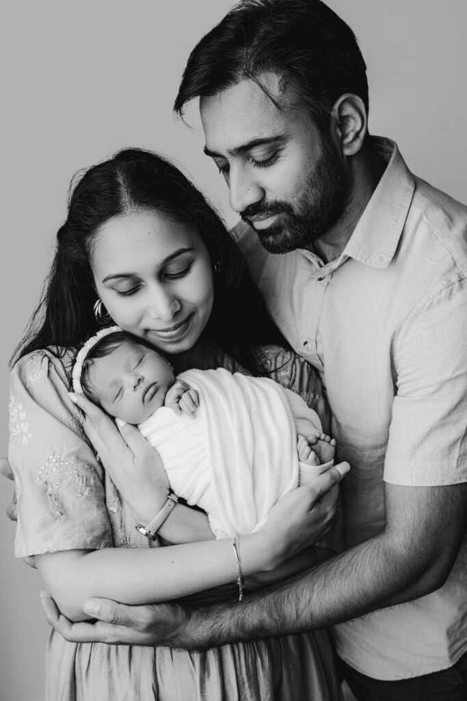 Black and white photo of mom and dad looking down while mom holds her rainbow baby close with love during Charlotte newborn photography session