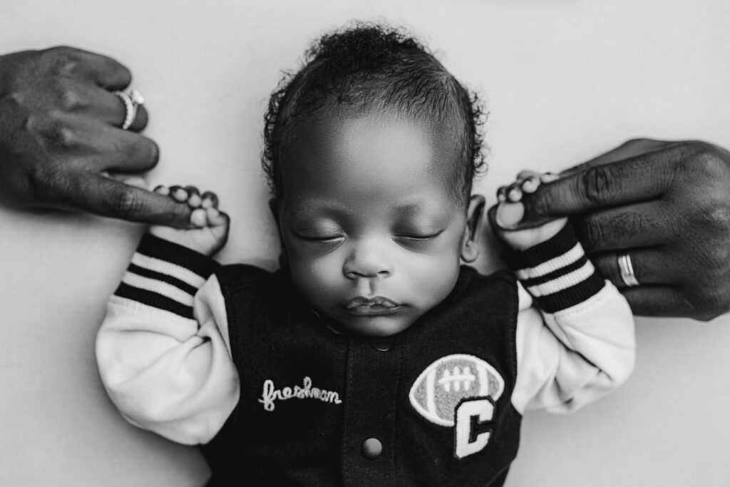 Black and white portrait of a premature newborn wearing a tiny varsity jacket, holding each parent’s hand, captured in a timeless style by Charlotte newborn photographer NicSo Studio.