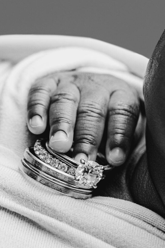 Black and white close-up of a newborn’s tiny fingers gently resting on their parents’ wedding rings