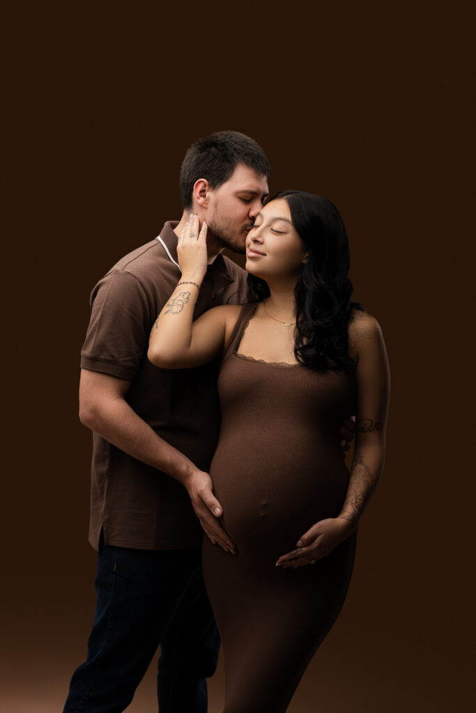 Expecting couple in matching brown tones sharing a tender kiss during a studio maternity session at NicSo Studio