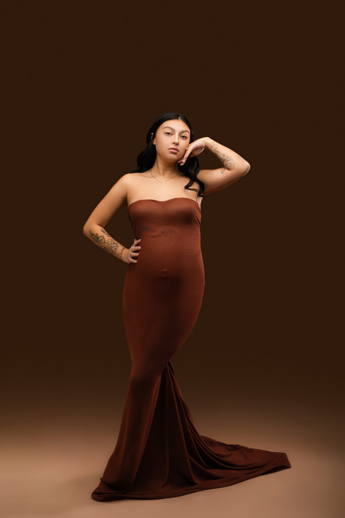  Expectant mother in a rust-colored strapless fitted gown with a long train, posing confidently during a studio maternity session at NicSo Studio in Waxhaw, NC