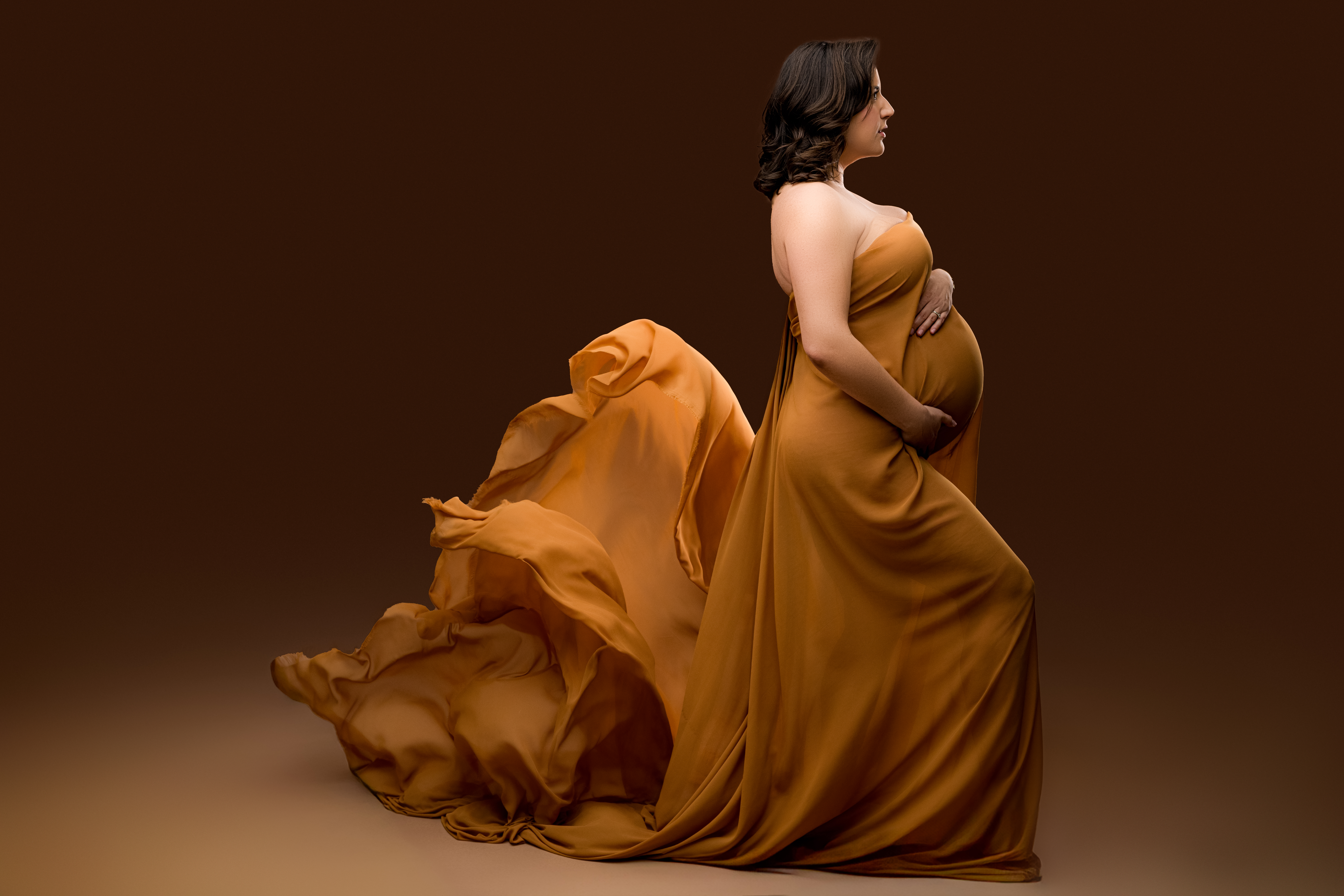 Pregnant woman in a flowing golden fabric wrap gown with dramatic movement, captured during a luxury studio maternity session at NicSo Studio near Charlotte, NC