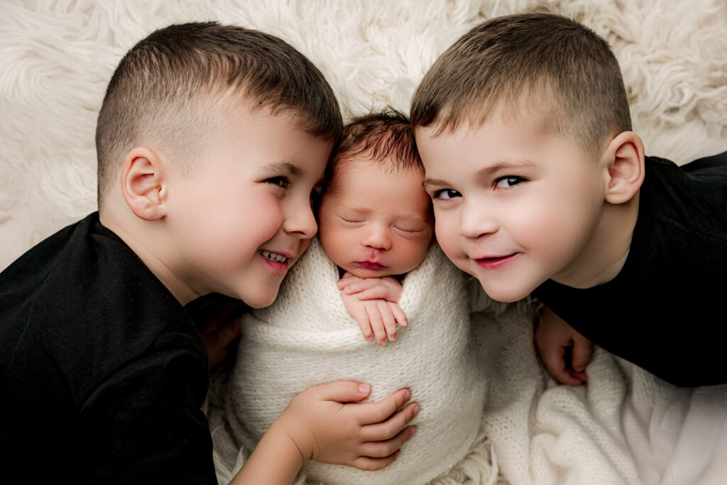 Two big brothers smiling as they hold their newborn baby brother during a sibling newborn session at NicSo Studio, Charlotte NC newborn photographer