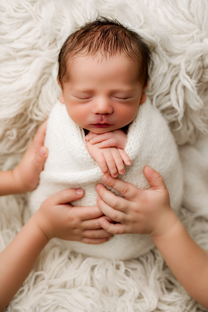 Big brothers gently holding their swaddled newborn baby brother with caring hands during a Charlotte newborn photography session at NicSo Studio