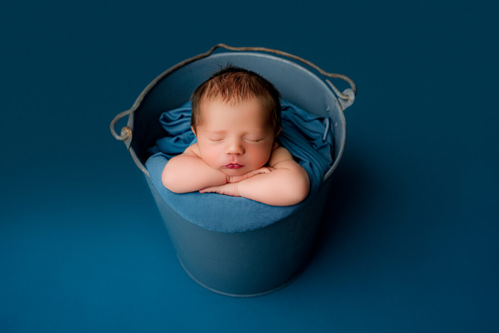 Newborn baby boy sleeping peacefully in a blue bucket prop during a studio newborn session at NicSo Studio.